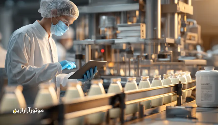 خط تولید لبنیات مدرن و تمیز با دستگاهها و نقالههای استیل در حال کار. تکنسین بهداشت با روپوش و تبلت در حال بررسی سطح روانکار دستگاهها است.