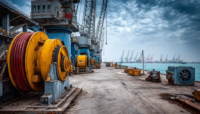 وینچ‌ها و تجهیزات بندری بزرگ در اسکله بندرعباس با پس‌زمینه دریا و جرثقیل‌ها | روغن صنعتی در بندرعباس