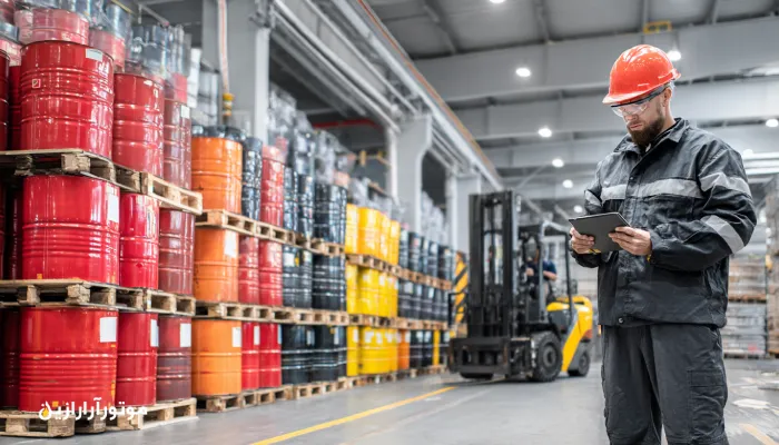 کارمند لجستیک و انبار موتورازین در حال مدیریت موجودی بشکه‌های روغن صنعتی با تبلت در انبار مرکزی