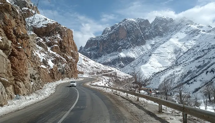 جاده کوهستانی برفی در مسیر اردبیل با خودروی سفید در حال عبور و چشم‌اندازی از کوه‌های پوشیده از برف