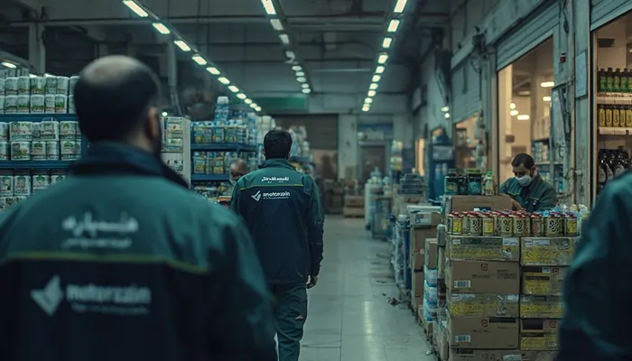 کارکنان موتورازین با لباس‌های کاری در حال بازدید از انبار بزرگ کالا و روانکارهای صنعتی در فضای بسته و روشن