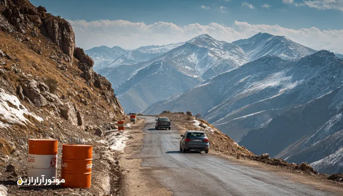 جاده کوهستانی کلاردشت–چالوس با خودروهای در حال صعود و بشکههای برند موتورازین در کنار جاده، نور روز واقعی و مناظر برفی. روغن موتور کلاردشت
